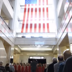 American flag handing from the ceiling of the Illinois State Library with people gathered below watching singers performing